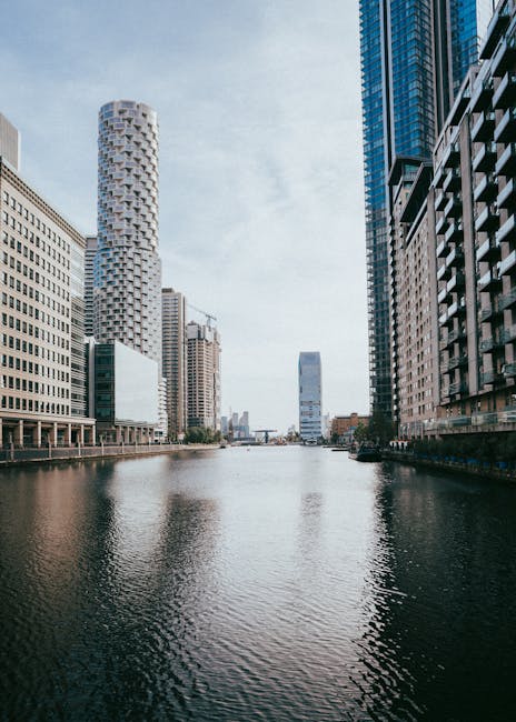 A wide view of the Canary Wharf area in London seen from across the waterway, with modern high-rise buildings on both sides of the river. The buildings include glass and steel structures, some with reflective surfaces, and one distinctive cylindrical tower with textured exterior. The sky is partly cloudy with patches of blue. The water in the foreground is calm, reflecting the buildings above. This urban landscape illustrates the central business district environment, typical of a professional setting where house removals and furniture transport services at Heron Quays apartment on Marsh Wall E14 are performed. The scene captures the scale and modern architecture characteristic of Canary Wharf, relevant to the context of property relocations managed by companies such as removalcompaniescanarywharf.co.uk, supporting the logistical aspects of packing, loading, and transporting household or office furniture within this commercial district.