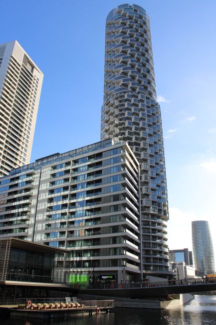 A tall, modern residential skyscraper in Canary Wharf with a distinctive cylindrical design featuring a pattern of staggered, protruding balconies arranged in a honeycomb-like structure. The building's facade is composed of glass and metal, reflecting the clear blue sky. In the foreground, there are lower-rise office and apartment buildings with large windows and multiple balconies, along with a bridge spanning a waterway. The scene is well-lit by natural sunlight, and the overall environment depicts an urban setting suitable for house removals and furniture transport services, as seen in professional moving activities and logistics in the Canary Wharf area.