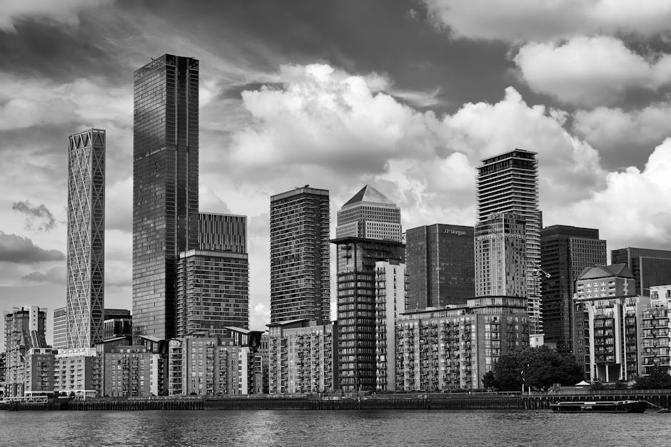 A wide view of the Canary Wharf area in London seen from across the waterway, with modern high-rise buildings on both sides of the river. The buildings include glass and steel structures, some with reflective surfaces, and one distinctive cylindrical tower with textured exterior. The sky is partly cloudy with patches of blue. The water in the foreground is calm, reflecting the buildings above. This urban landscape illustrates the central business district environment, typical of a professional setting where house removals and furniture transport services at Heron Quays apartment on Marsh Wall E14 are performed. The scene captures the scale and modern architecture characteristic of Canary Wharf, relevant to the context of property relocations managed by companies such as removalcompaniescanarywharf.co.uk, supporting the logistical aspects of packing, loading, and transporting household or office furniture within this commercial district.