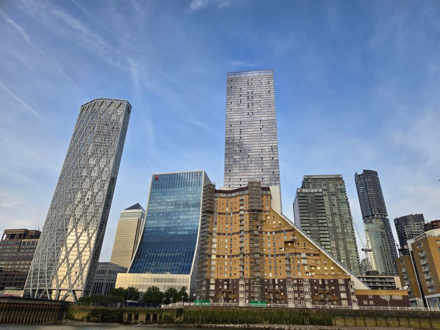 A cityscape view of Canary Wharf, featuring several modern high-rise buildings with glass facades under a clear blue sky. In the foreground, a street-level scene shows a residential building with a unique sloped roof covered in brown and beige facade panels, surrounded by a pavement and some trees. To the left, a distinctive office tower with a lattice external structure is visible, while other skyscrapers with varying heights and architectural styles fill the background. The scene captures the urban environment where home relocation and furniture transport services, such as those provided by [COMPANY_NAME], may operate during a house move, with loading and moving processes occurring within this high-rise setting.