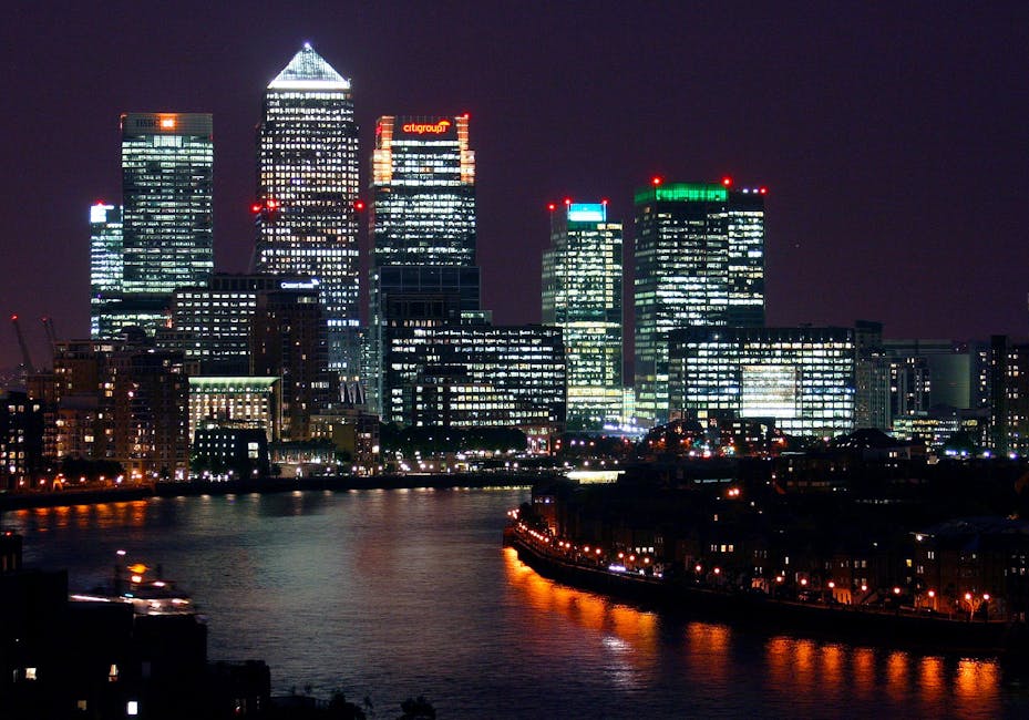 A nighttime cityscape of Canary Wharf with illuminated skyscrapers along the river Thames. The buildings include modern glass office towers with bright interior lights, some displaying company logos such as Citigroup. The river reflects the city lights, and in the foreground, a paved area with streetlights suggests an area where moving trucks or vans may be parked. The scene indicates an urban environment suitable for home relocation and furniture transport, with visible logistics such as loading or unloading activities possibly occurring nearby, supported by companies like [COMPANY_NAME] involved in house removals and packing during the moving process.