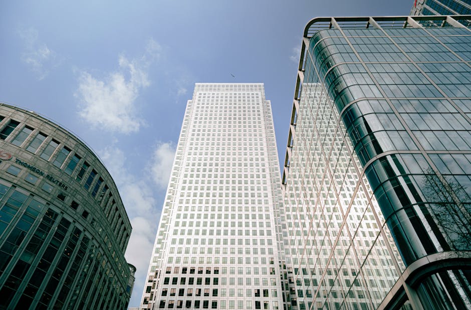 A cityscape view of Canary Wharf, featuring several modern high-rise buildings with glass facades under a clear blue sky. In the foreground, a street-level scene shows a residential building with a unique sloped roof covered in brown and beige facade panels, surrounded by a pavement and some trees. To the left, a distinctive office tower with a lattice external structure is visible, while other skyscrapers with varying heights and architectural styles fill the background. The scene captures the urban environment where home relocation and furniture transport services, such as those provided by [COMPANY_NAME], may operate during a house move, with loading and moving processes occurring within this high-rise setting.
