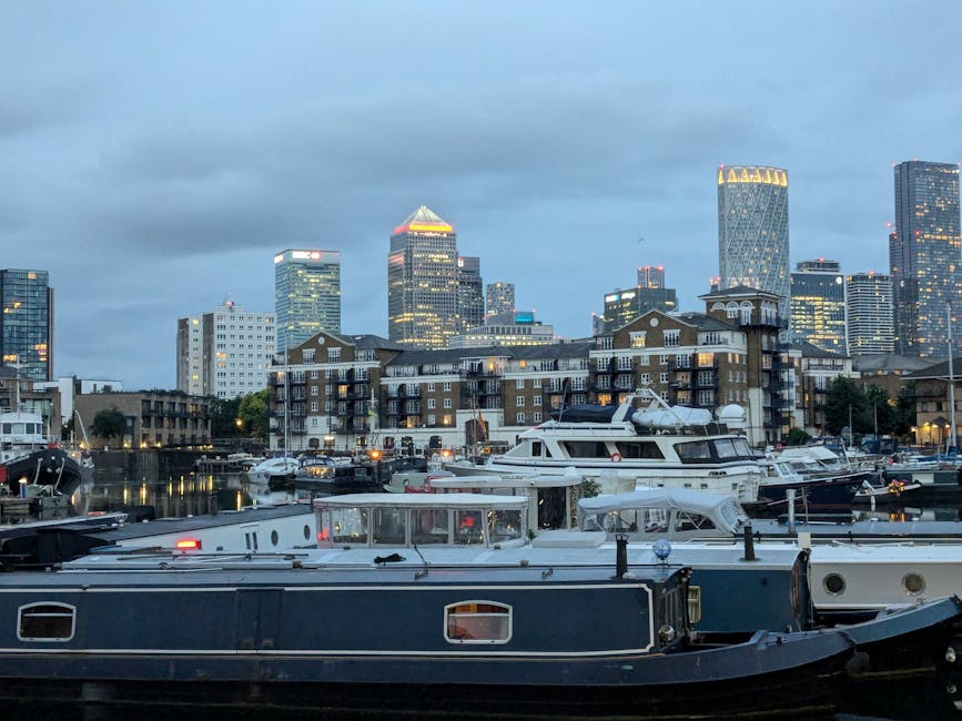 A view of Marina Quays, a residential waterfront area in Canary Wharf, with multiple modern apartment buildings featuring brick facades, white balconies, and large windows. The marina is filled with several moored boats, including luxury yachts and smaller vessels, some covered in protective tarps or plastic wrapping. In the foreground, a narrow walkway along the water's edge is visible, with a few bollards and mooring posts. Across the water, the skyline displays tall office buildings and skyscrapers illuminated by interior and exterior lighting, with reflective glass facades and distinctive architectural designs. The sky appears overcast, casting soft, diffused light over the scene. This setting is associated with home relocation and furniture transport services, illustrating the urban environment where [COMPANY_NAME] offers removals and moving logistics in the West India Quay to Canary Wharf area, as part of professional packing and loading processes observed nearby.
