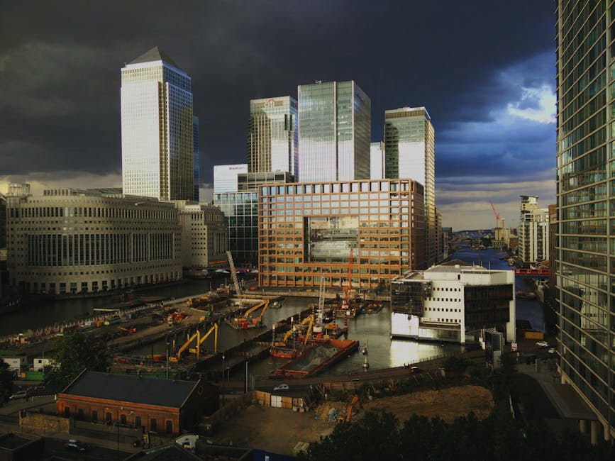 A daytime view of an urban waterfront area with several high-rise office buildings and modern glass towers. In the foreground, construction equipment including cranes and excavators are positioned along the water’s edge, with some machinery actively engaged in lifting or moving materials. There are stacks of wooden pallets and piles of construction materials, as well as a few parked vehicles and truck trailers nearby. The surrounding environment features a mix of commercial and government buildings, with partly cloudy skies overhead casting shadows over the scene. This setting depicts a home relocation or furniture transport process supported by professional removals services, as part of an ongoing building development or renovation project in the West India Quay to Canary Wharf estate area.